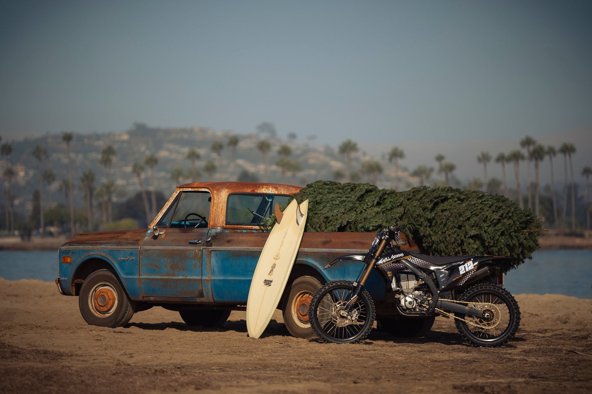 Rustic truck with a surfboard and motorcycle on a beach with palm trees in the background