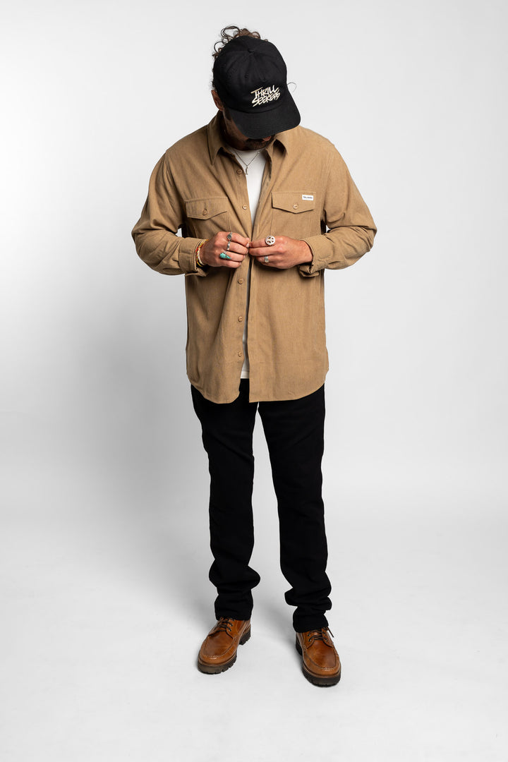 Man wearing a khaki button up long sleeve, black pants, and a black cap on a white background