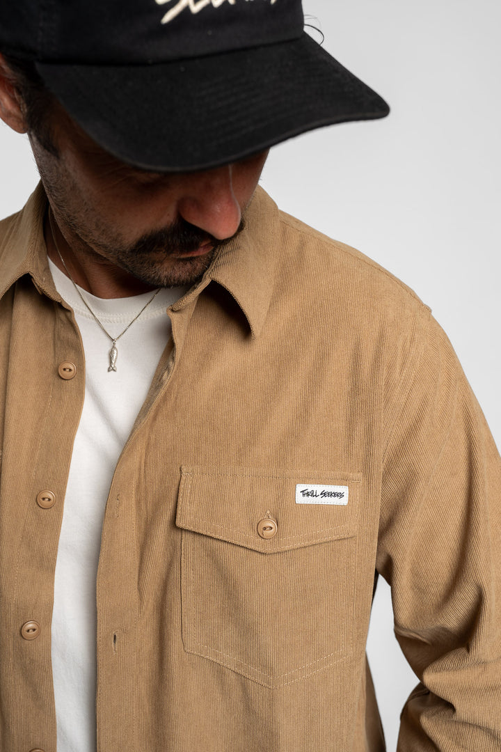 Man wearing a khaki long sleeve button up shirt with a Thrill Seekers logo and a black cap.