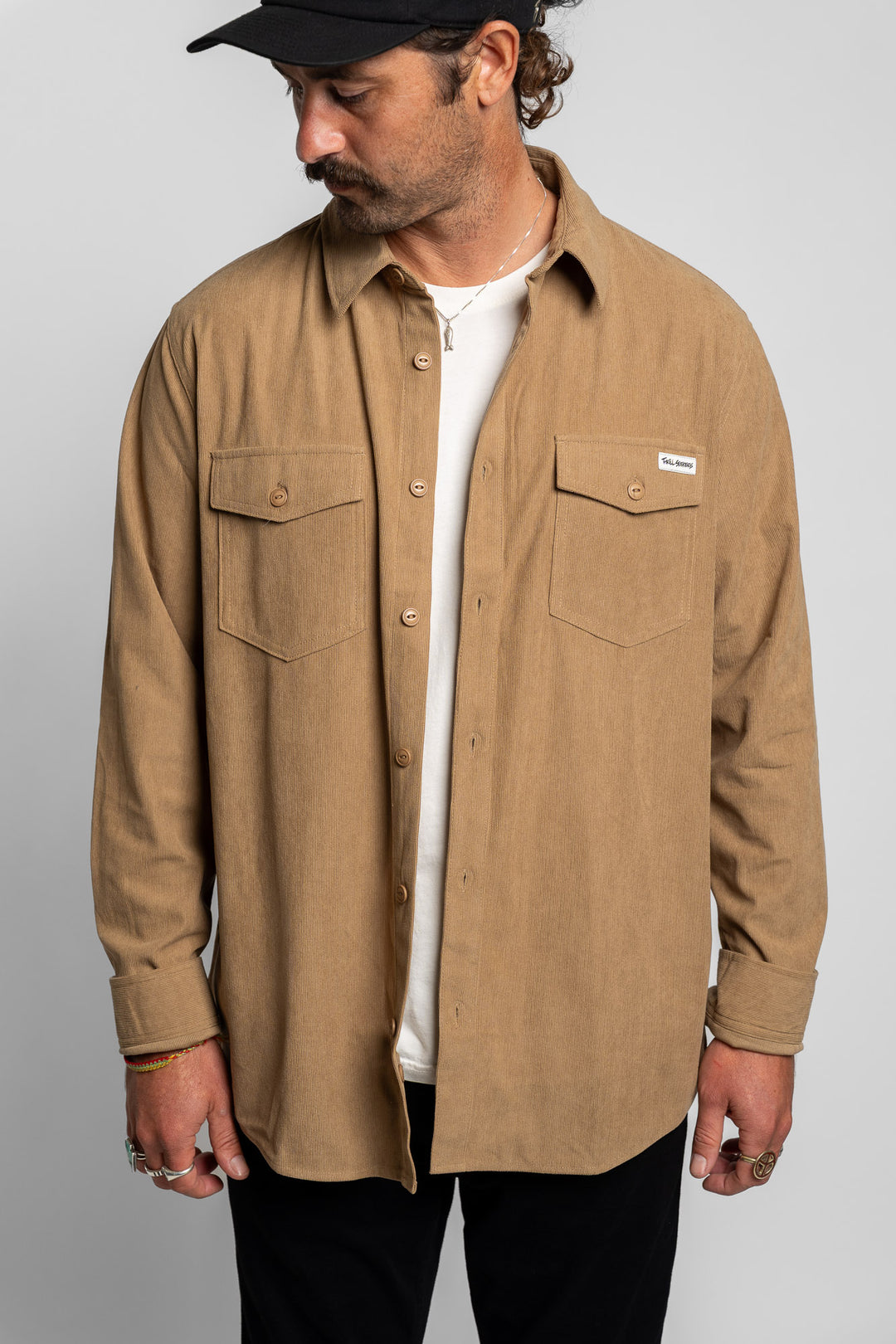 Man wearing a khaki long sleeve button up shirt over a white t-shirt with a plain background