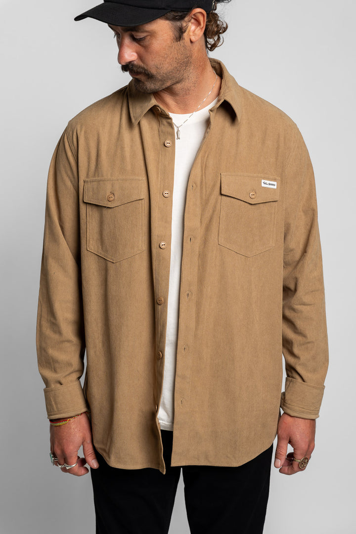 Man wearing a khaki long sleeve button up shirt over a white t-shirt with a plain background
