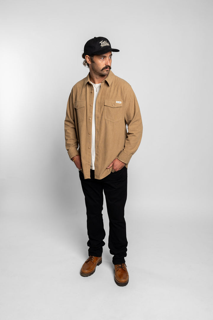 Man wearing a khaki button up long sleeve, black pants, and a black cap on a white background