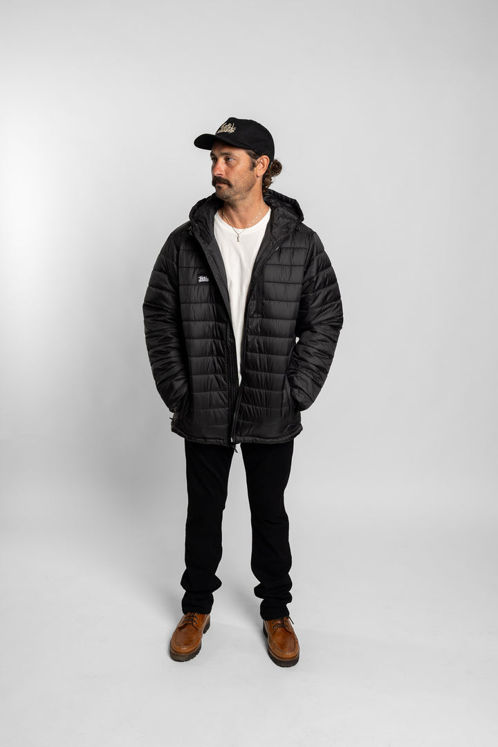 Man wearing a black puffer jacket, white shirt, black pants, and brown boots on a white background