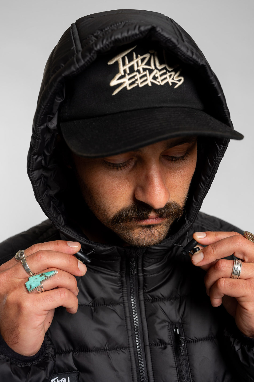 Person wearing a black puffer jacket hood and cap with 'Thrill Seekers' logo on a white background