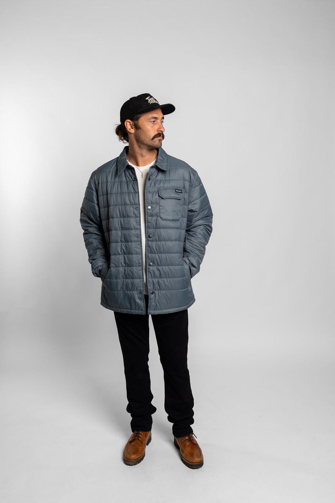 Man wearing a blue quilted jacket, black cap, and brown boots on a white background