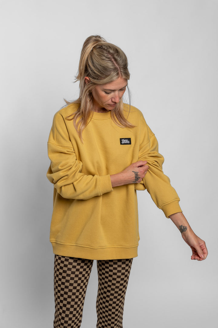 Person wearing a mustard yellow sweatshirt with a small logo on a plain background