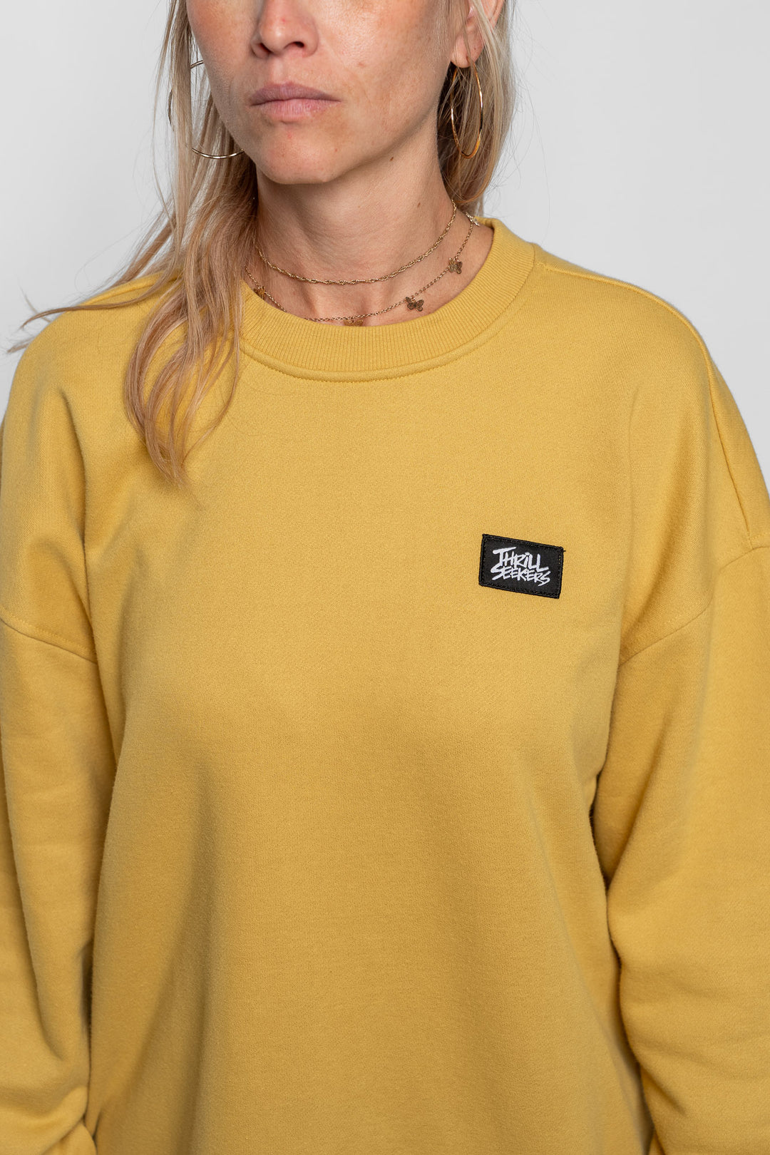 Person wearing a mustard yellow sweatshirt with a small logo on a plain background