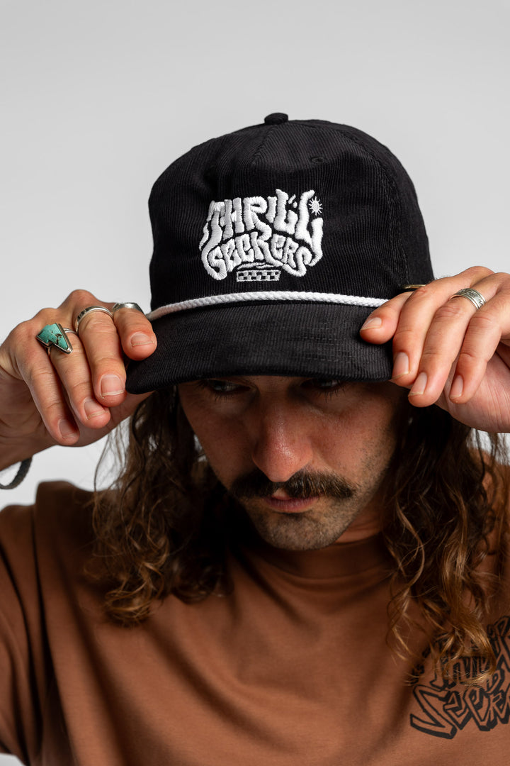 Person wearing a black corduroy cap with 'Thrill Seekers' logo on a white background