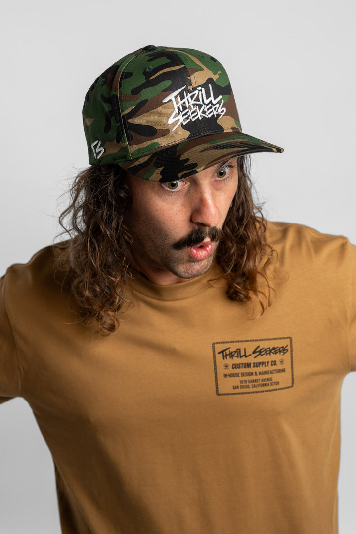 Person wearing a camo snapback hat with 'Thrill Seekers' logo in white