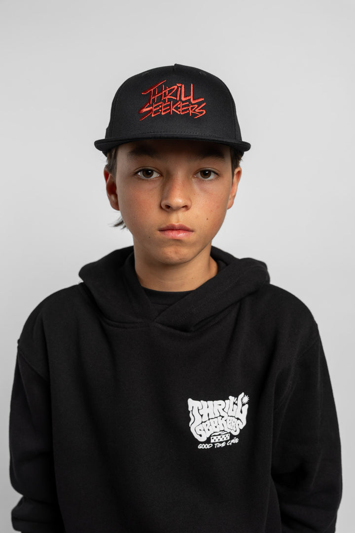 Kid wearing black cap with cardinal red 'Thrill Seekers' logo on a white background