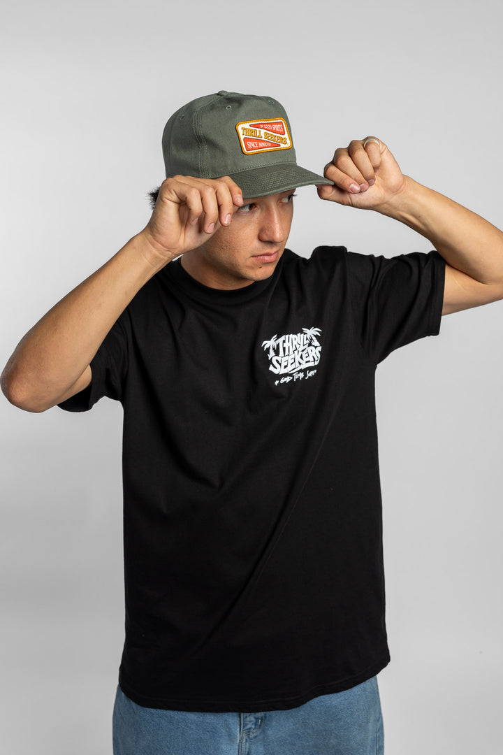 Person wearing a black t-shirt with a green 'Thrill Seekers' logo cap on a white background